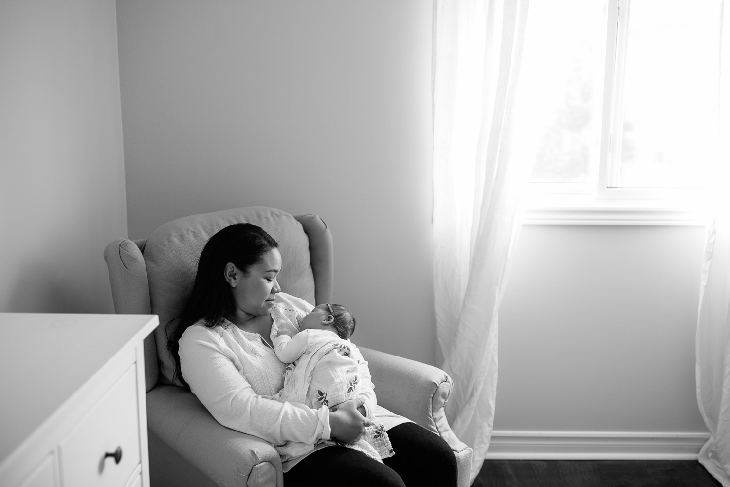 mom with long dark hair in white shirt sitting in nursery glider chair with 2 week old baby girl in onesie and swaddle sleeping on mother's chest - Markham Lifestyle Photography