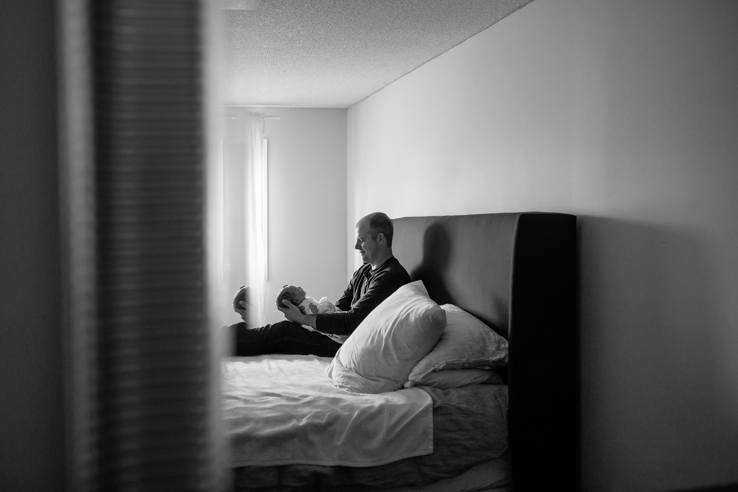 dad sitting on master bed holding 2 week old baby girl in his lap, smiling at her, reflection in mirror - Newmarket In-Home Photography