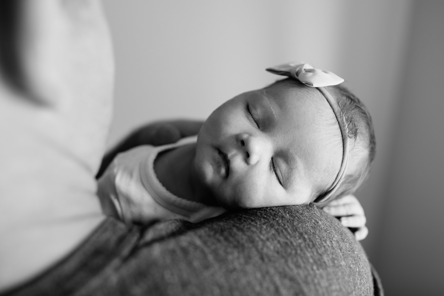 dad sitting on edge of bed holding 2 week old baby girl wearing bow headband, sleeping on father's shoulder - Barrie In-Home Photography
