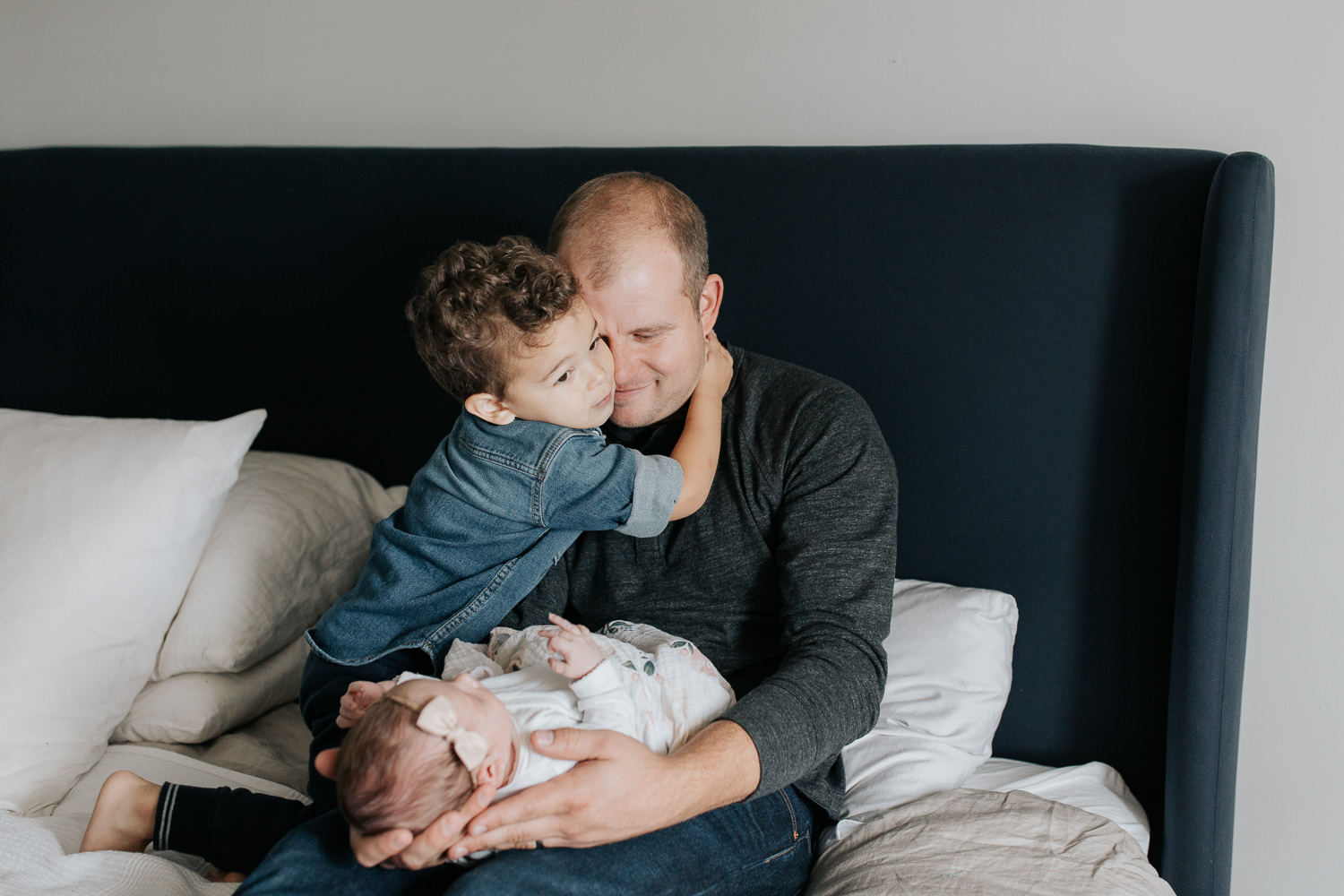 dad sitting on master bed holding 2 week old baby girl in his lap, 2 year old son hugging father around neck - Stouffville In-Home Photos