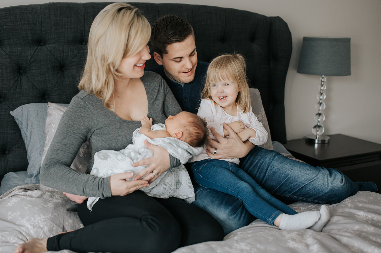  family of four sitting on bed, mom holding 2 week old baby boy 2 year old girl sitting on dad's lap smiling - Markham Lifestyle Photography