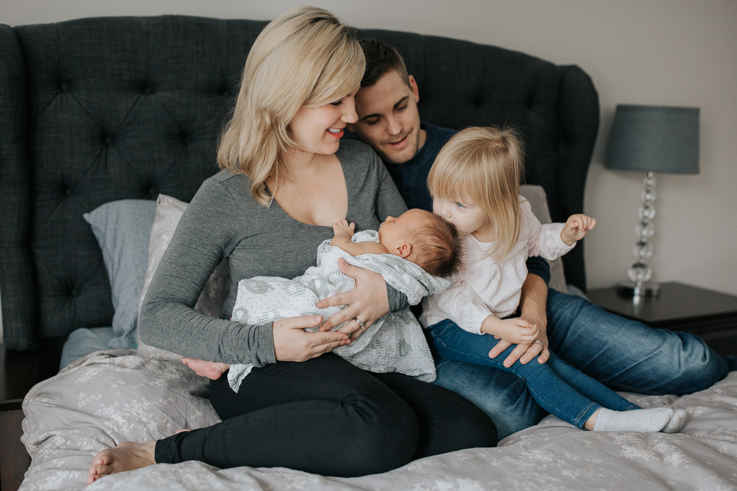  family of four sitting on bed, mom holding 2 week old baby boy 2 year old girl sitting on dad's lap kissing brother's head - Barrie Lifestyle Photography