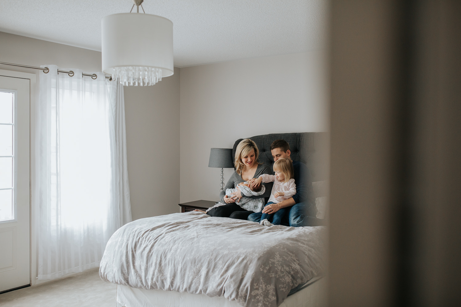  family of four sitting on bed, mom holding 2 week old baby boy 2 year old girl sitting on dad's lap touching brother's head - Newmarket Lifestyle Photography