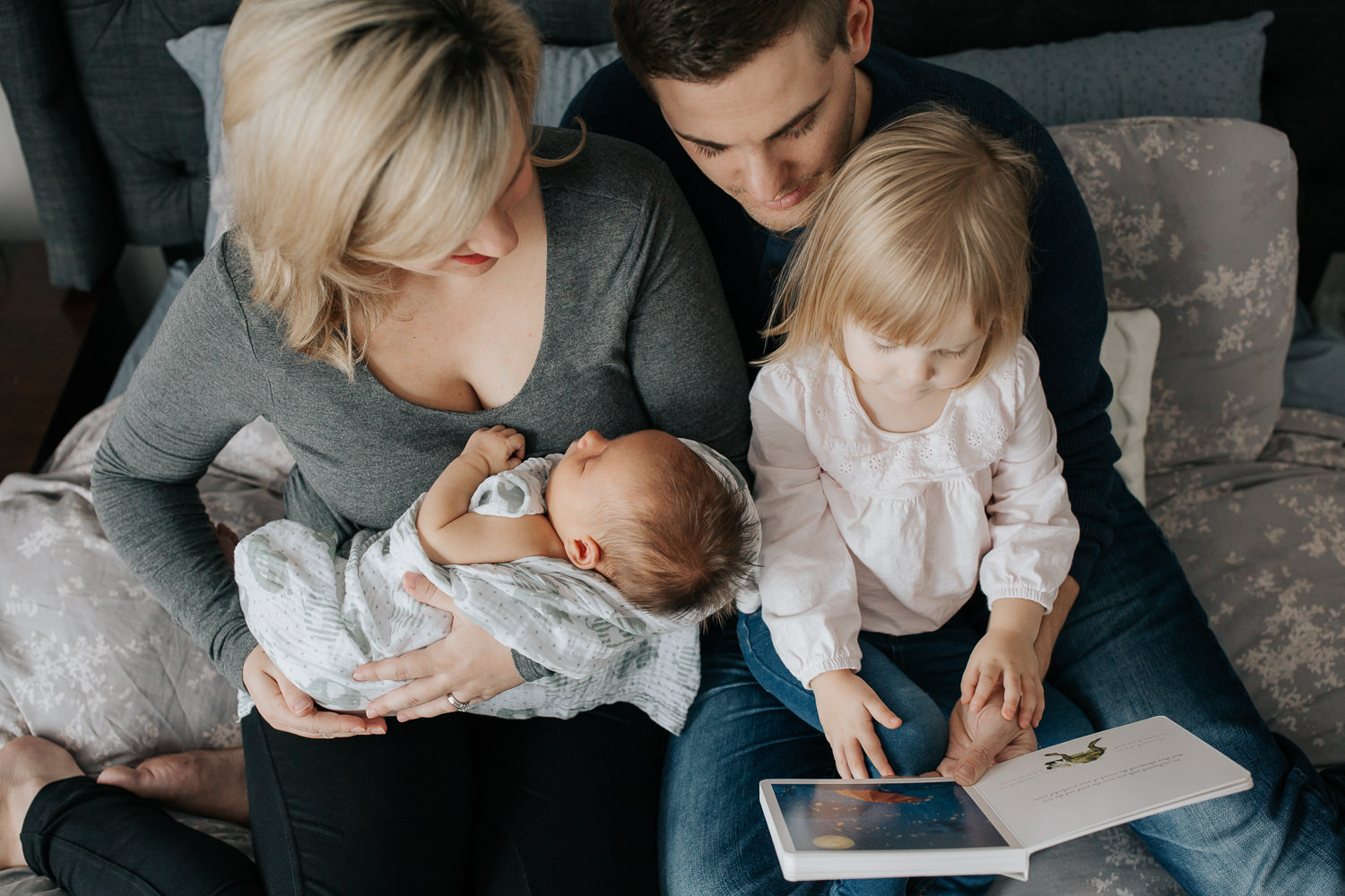  family of four sitting on bed, mom holding 2 week old baby boy 2 year old girl sitting on dad's lap reading story book - GTA Lifestyle Photography