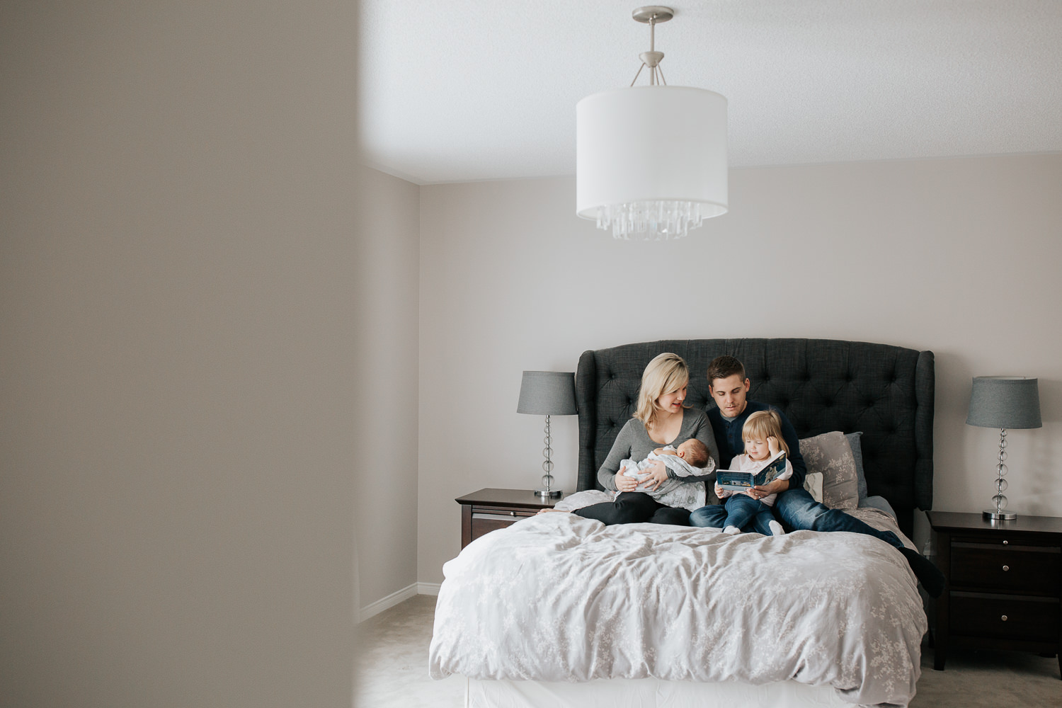  family of four sitting on bed, mom holding 2 week old baby boy 2 year old girl sitting on dad's lap reading story book - York Region Lifestyle Photography