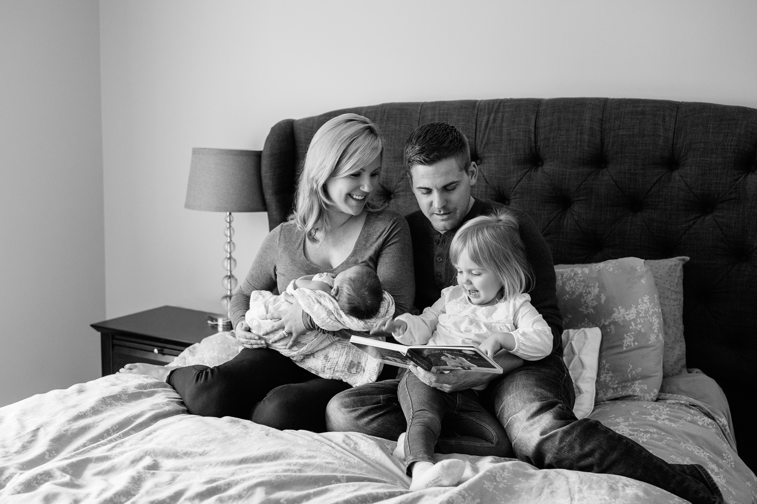  family of four sitting on bed, mom holding 2 week old baby boy 2 year old girl sitting on dad's lap reading story book - Stouffville Lifestyle Photography