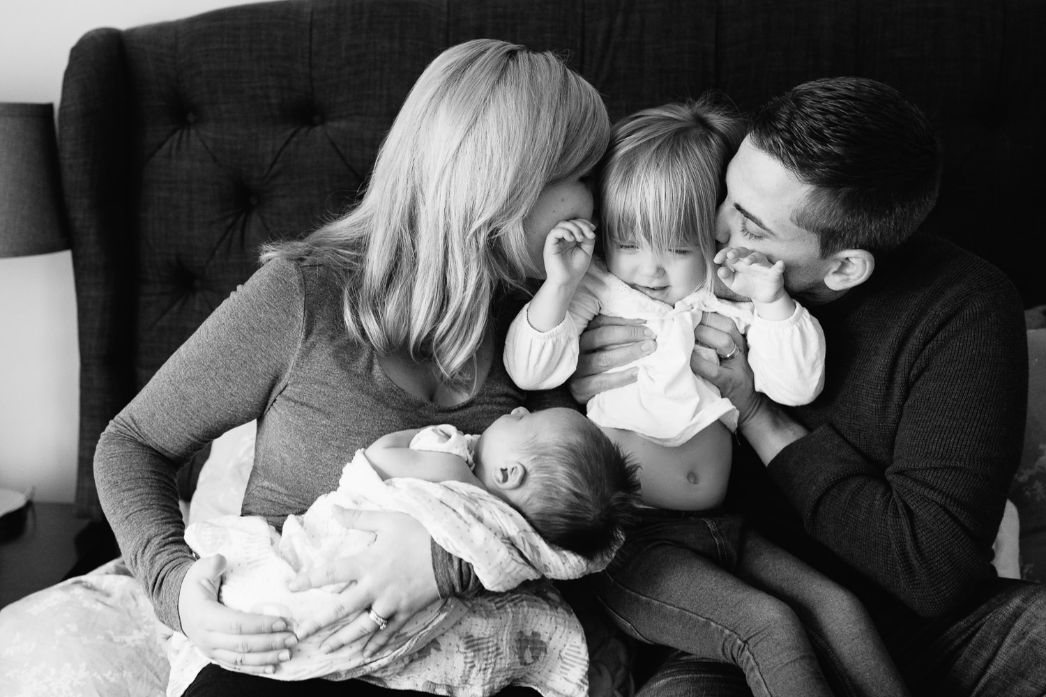  family of four sitting on bed, mom holding 2 week old baby boy 2 year old girl sitting on dad's lap, parents kissing daughter's cheeks - Newmarket Lifestyle Photography