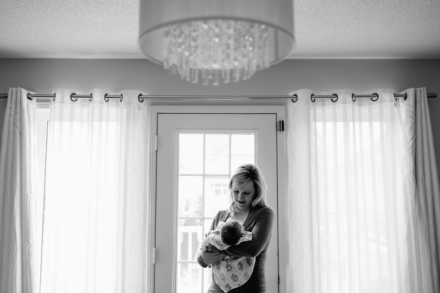 Barrie In-Home Photos - blonde mom adoring her new 2 week old baby boy standing by the door with a crystal chandelier up above
