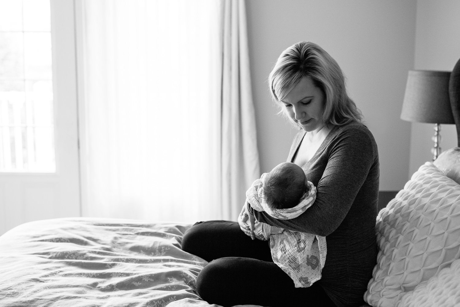 mother sitting on master bed holding 2 week old baby boy in swaddle in her arms, mom looking down at son - Barrie Lifestyle Photos