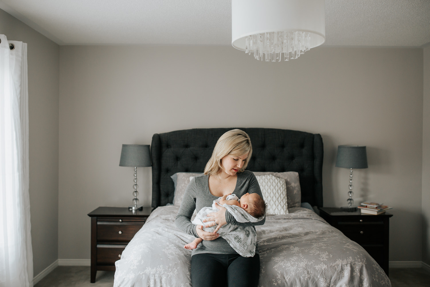 mother sitting on edge of master bed holding 2 week old baby boy in swaddle in her arms, mom looking down at son - GTA Lifestyle Photos