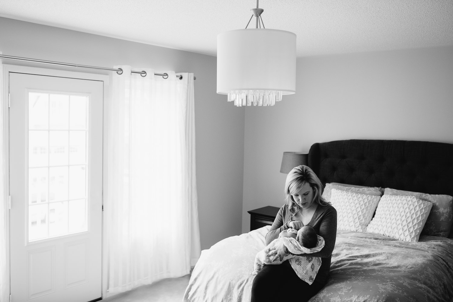 mother sitting on edge of master bed holding 2 week old baby boy in swaddle in her arms, mom looking down at son - Stouffville Lifestyle Photography 