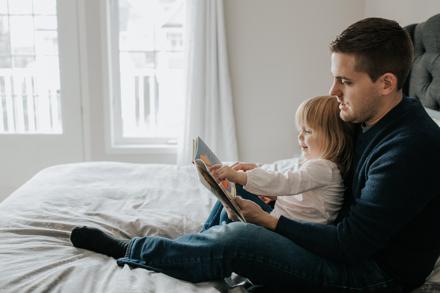 2 year old toddler girl sitting in father's lap on master bed reading a story book, pointing to page smiling - GTA In-Home Photos