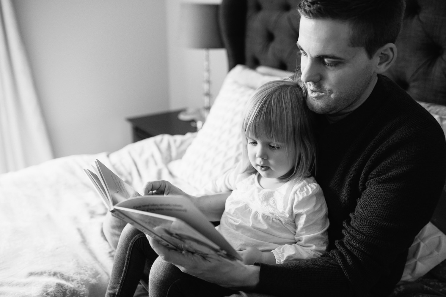 2 year old toddler girl sitting in father's lap on master bed reading a story book - York Region In-Home Photos 