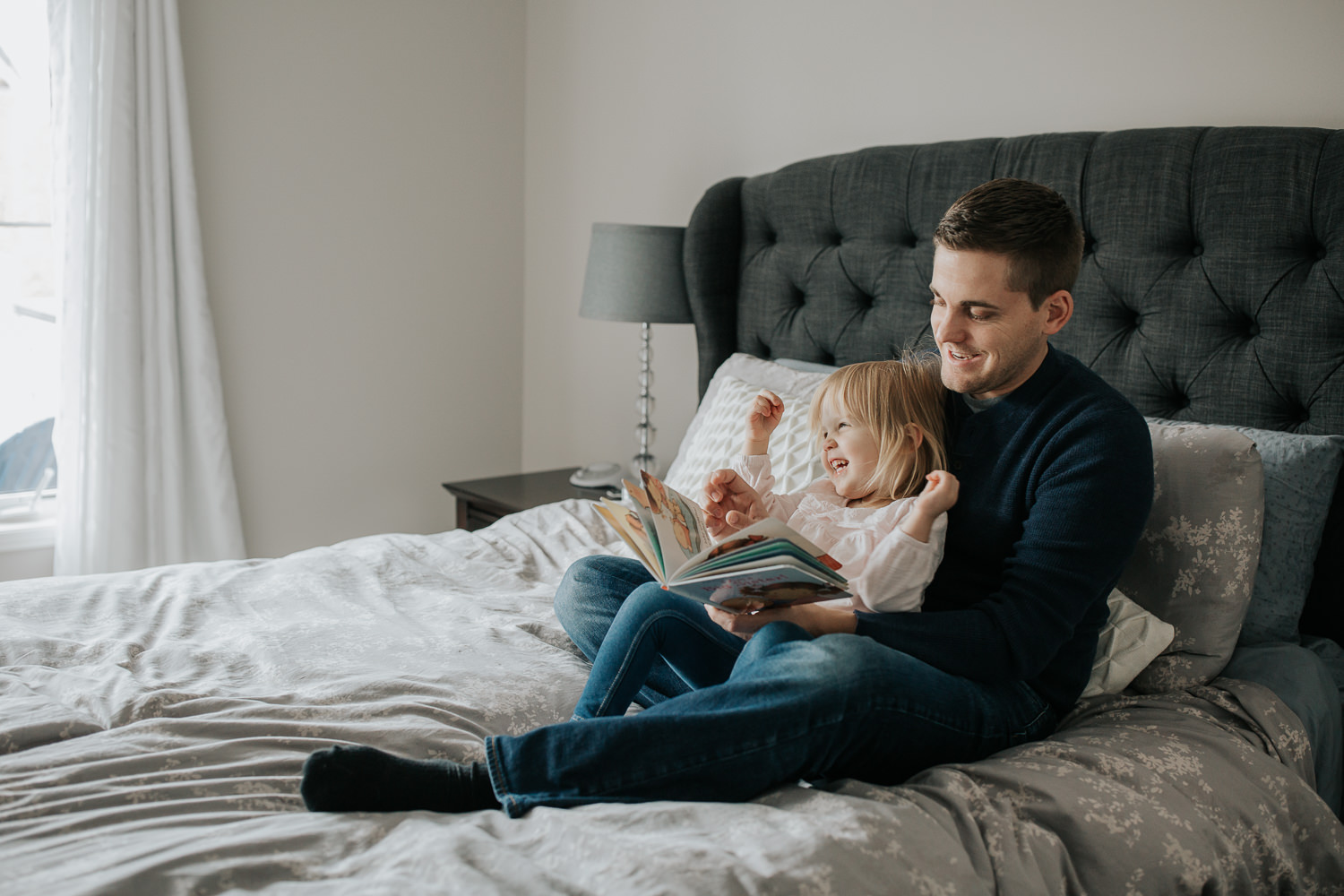 2 year old toddler girl sitting in father's lap on master bed reading a story book, daughter with hands in air laughing - Markham Lifestyle Photos