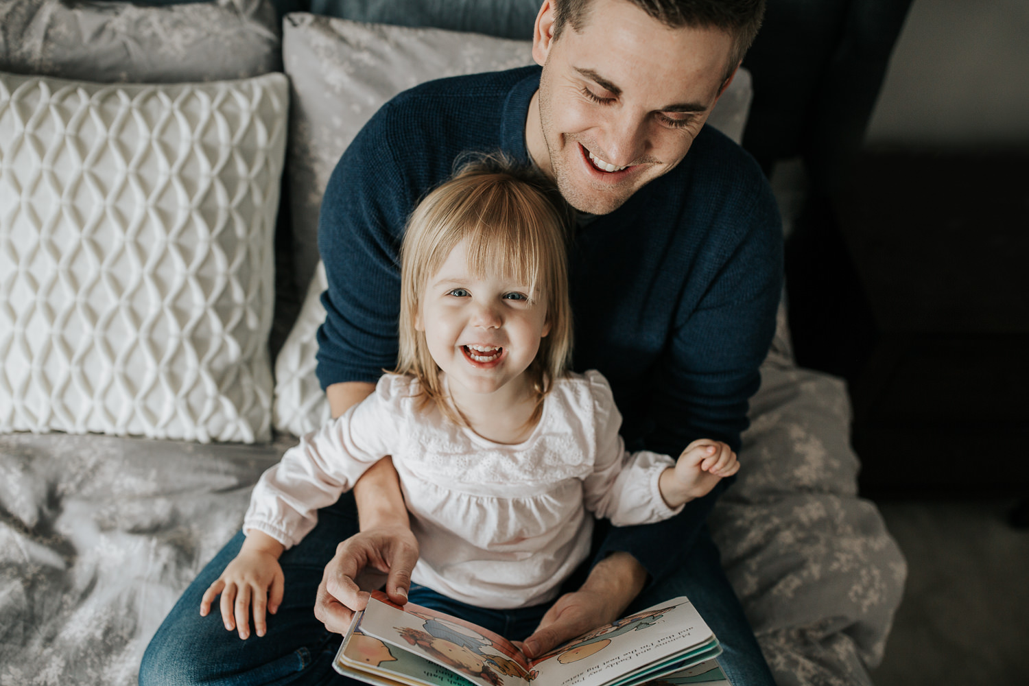 2 year old toddler girl sitting in father's lap on master bed reading a story book, daughter smiling up at camera - Newmarket Lifestyle Photos