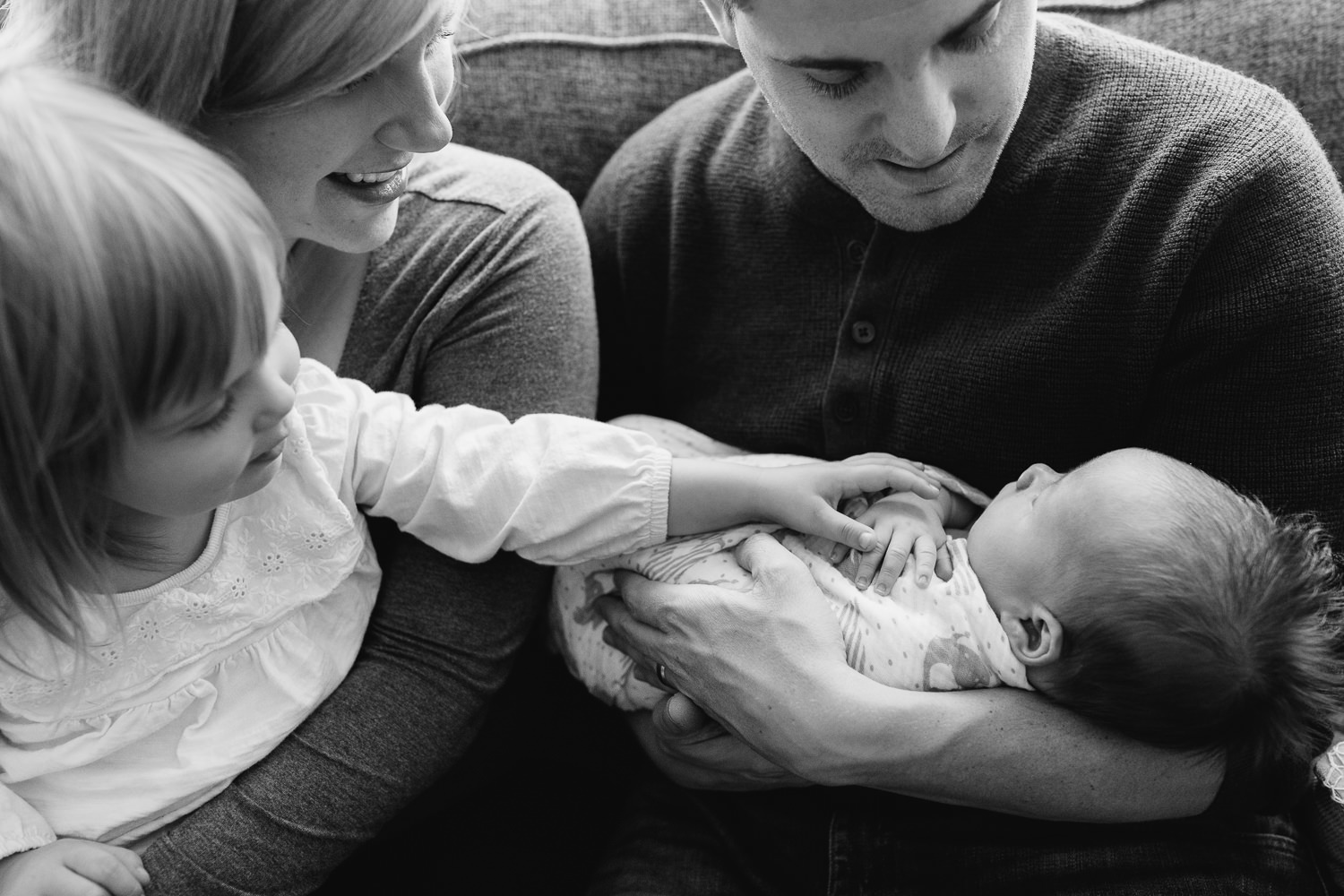 - family of four smiling together on a living room couch, 2 week old baby boy in dad's arms, toddler girl in mom's lap, big sister holding little brother's hand - Barrie Lifestyle Photos 