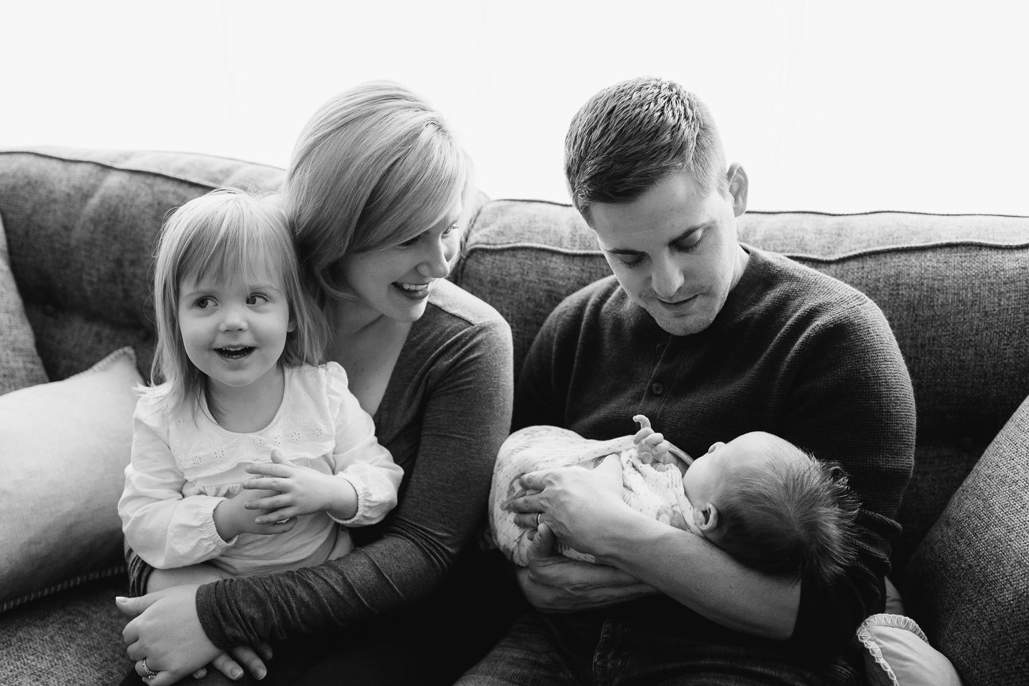 Stouffville Lifestyle Photos - family of four smiling together on a living room couch, 2 week old baby boy in dad's arms, toddler girl in mom's lap, parents smiling at son