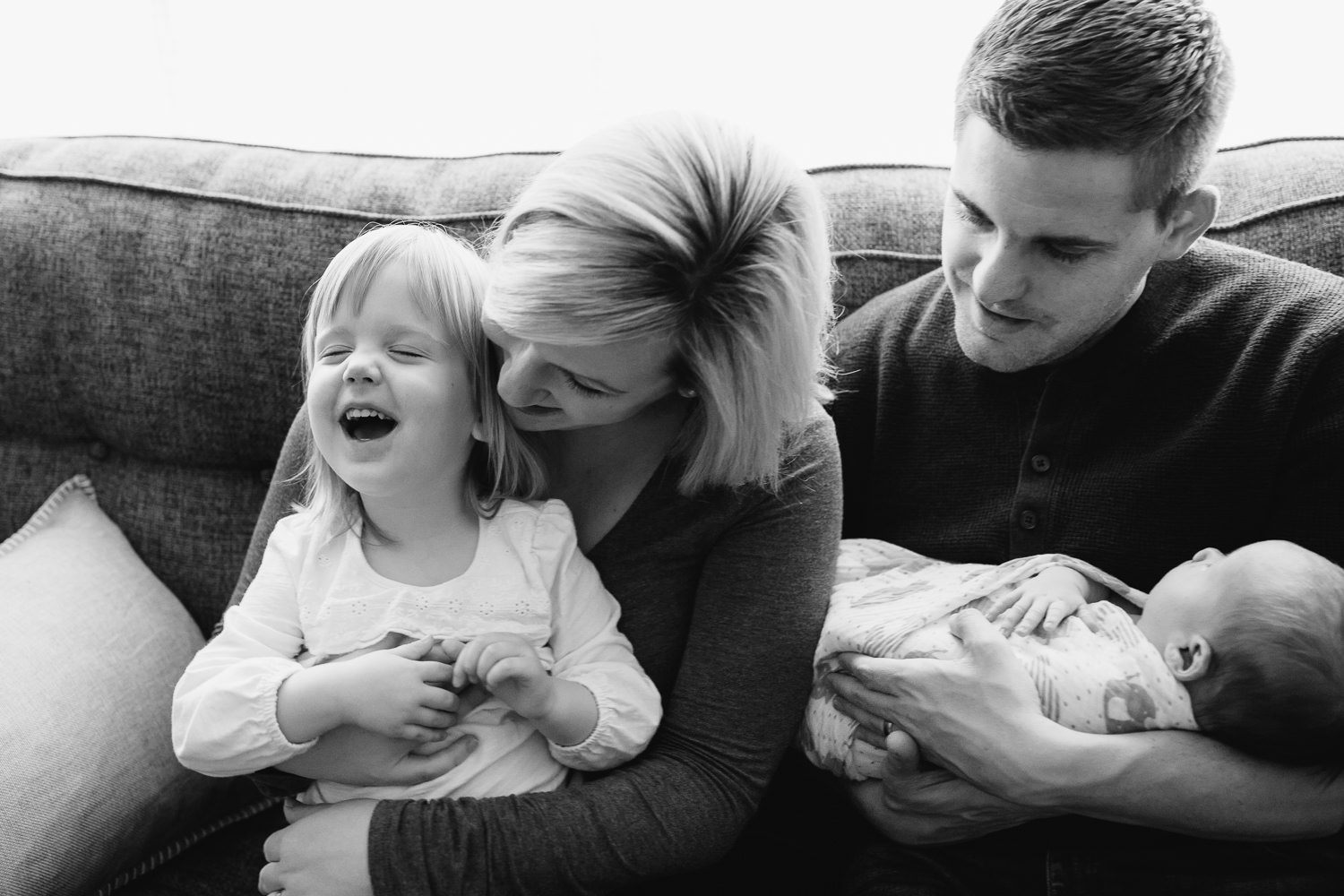 - family of four smiling together on a living room couch, 2 week old baby boy in dad's arms, toddler girl in mom's lap, daughter laughing as mother hugs her - Newmarket Lifestyle Photography 