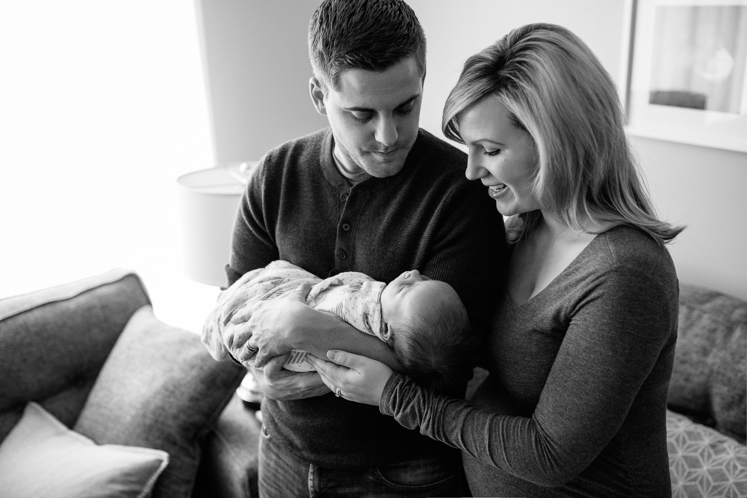 husband and wife standing in living room, father holding 2 week old baby boy in his arms, mom's hand on son - York Region Lifestyle Photography