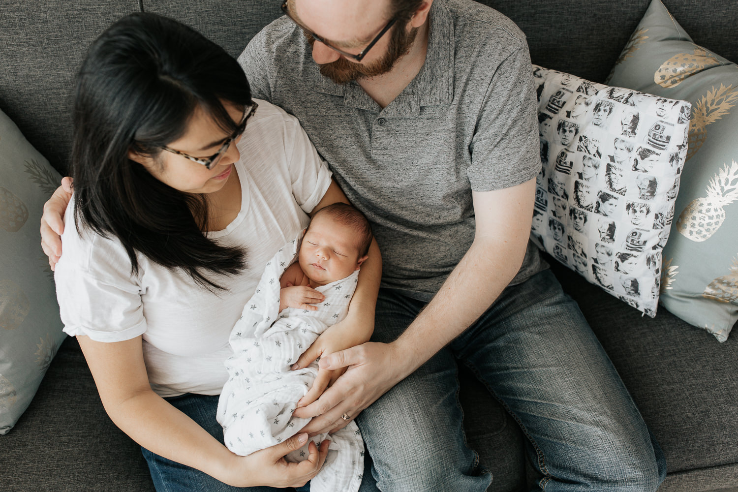 family of 3 sitting on couch, new mom holding sleeping, swaddled 2 week old baby boy, dad next to them with arm around wife and hand on son, parents smiling - GTA Lifestyle Photography