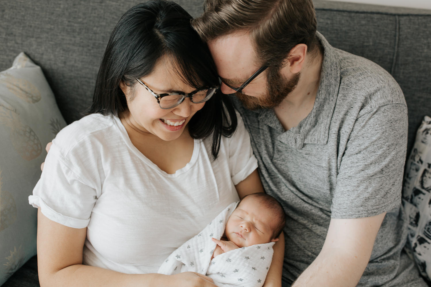 family of 3 sitting on couch, new mom holding sleeping, swaddled 2 week old baby boy, dad next to them leaning head against wife's and hand on son - Newmarket In-Home Photography