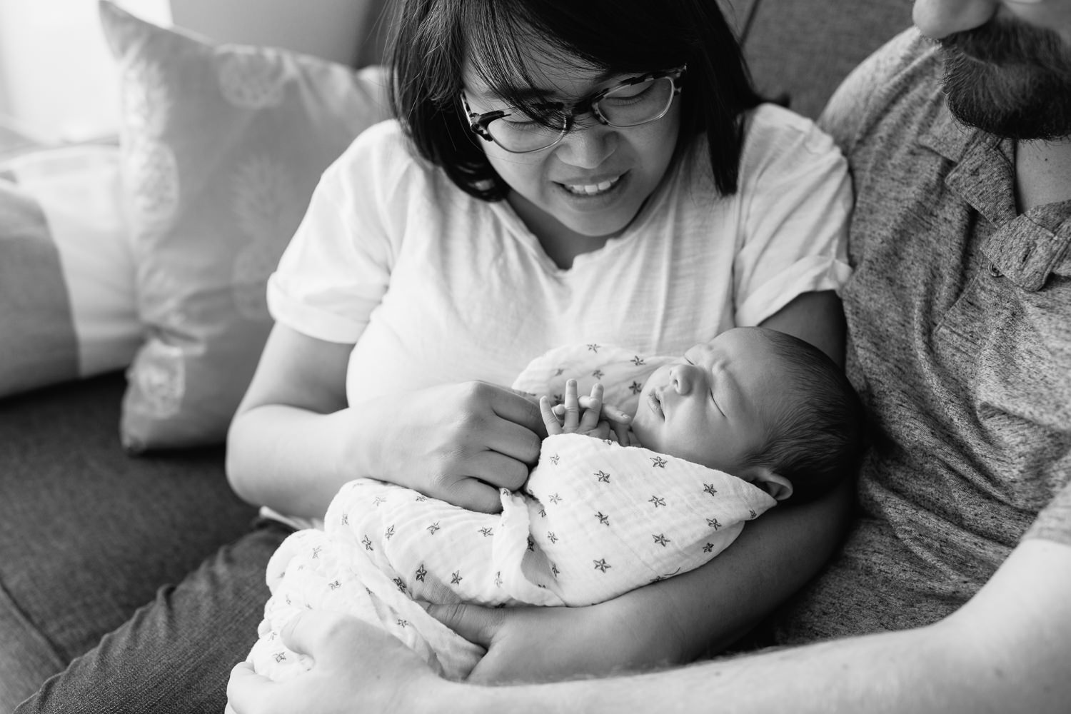 family of 3 sitting on couch, new mom holding sleeping, swaddled 2 week old baby boy, dad next to them with arm around wife and hand on son, parents smiling - York Region Lifestyle Photography