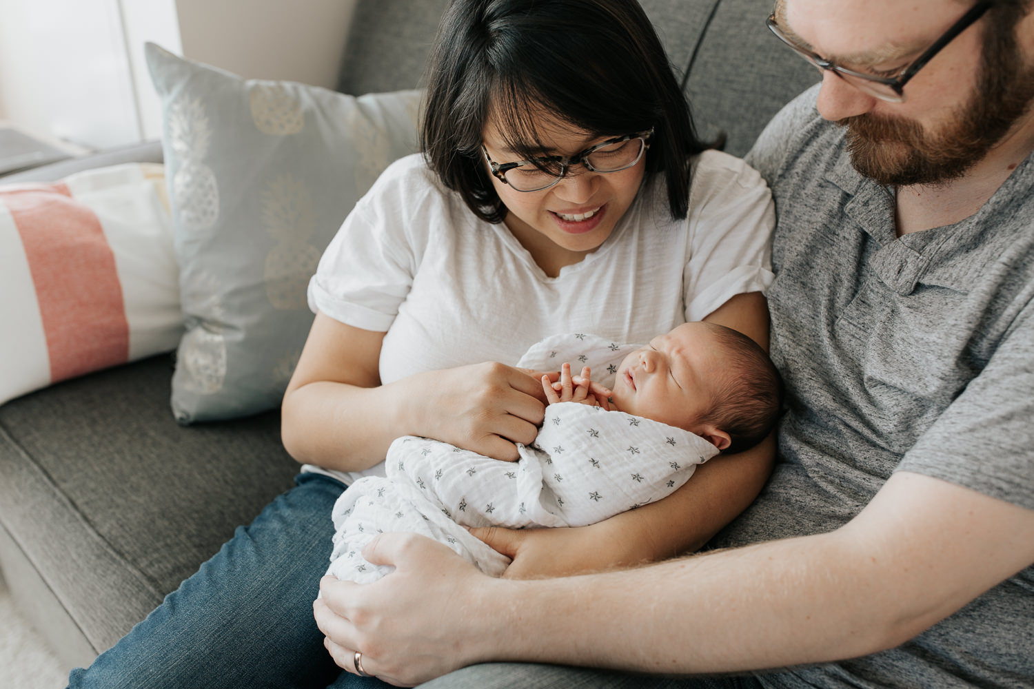 family of 3 sitting on couch, new mom holding sleeping, swaddled 2 week old baby boy, dad next to them with arm around wife and hand on son, parents smiling - Newmarket Lifestyle Photography