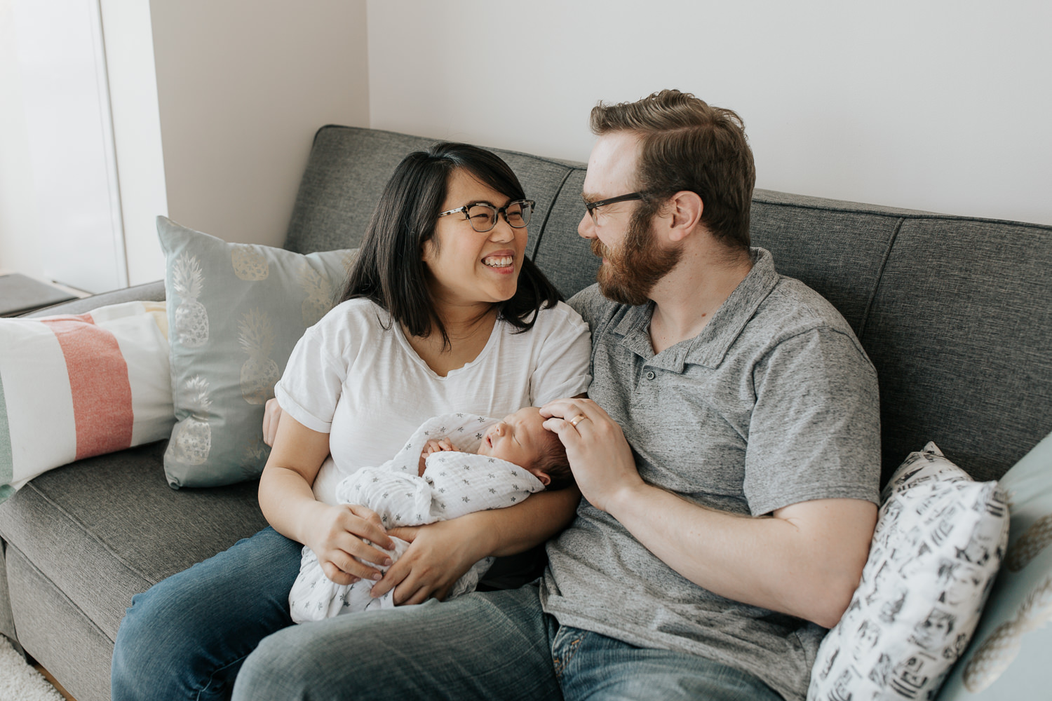 family of 3 sitting on couch, new mom holding sleeping, swaddled 2 week old baby boy, dad next to them with hand on son's head, parents smiling at one another - Markham In-Home Photography