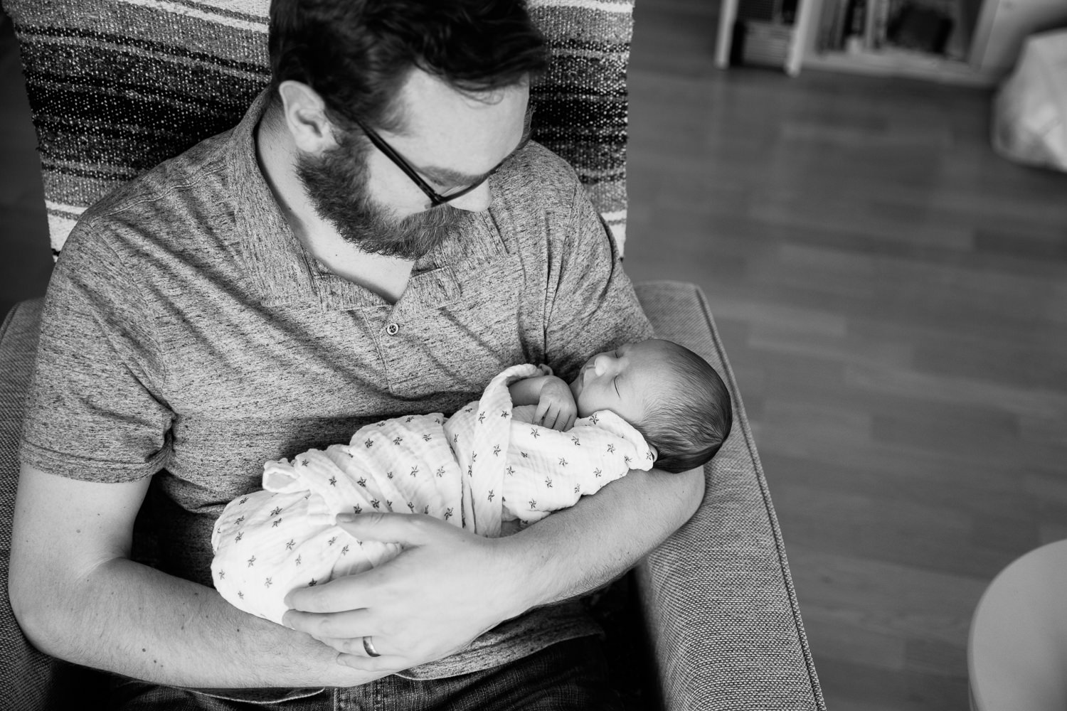 new father sitting in chair holding 2 week old baby boy in swaddle with dark hair, sleeping with hand near chin - Barrie In-Home Photography