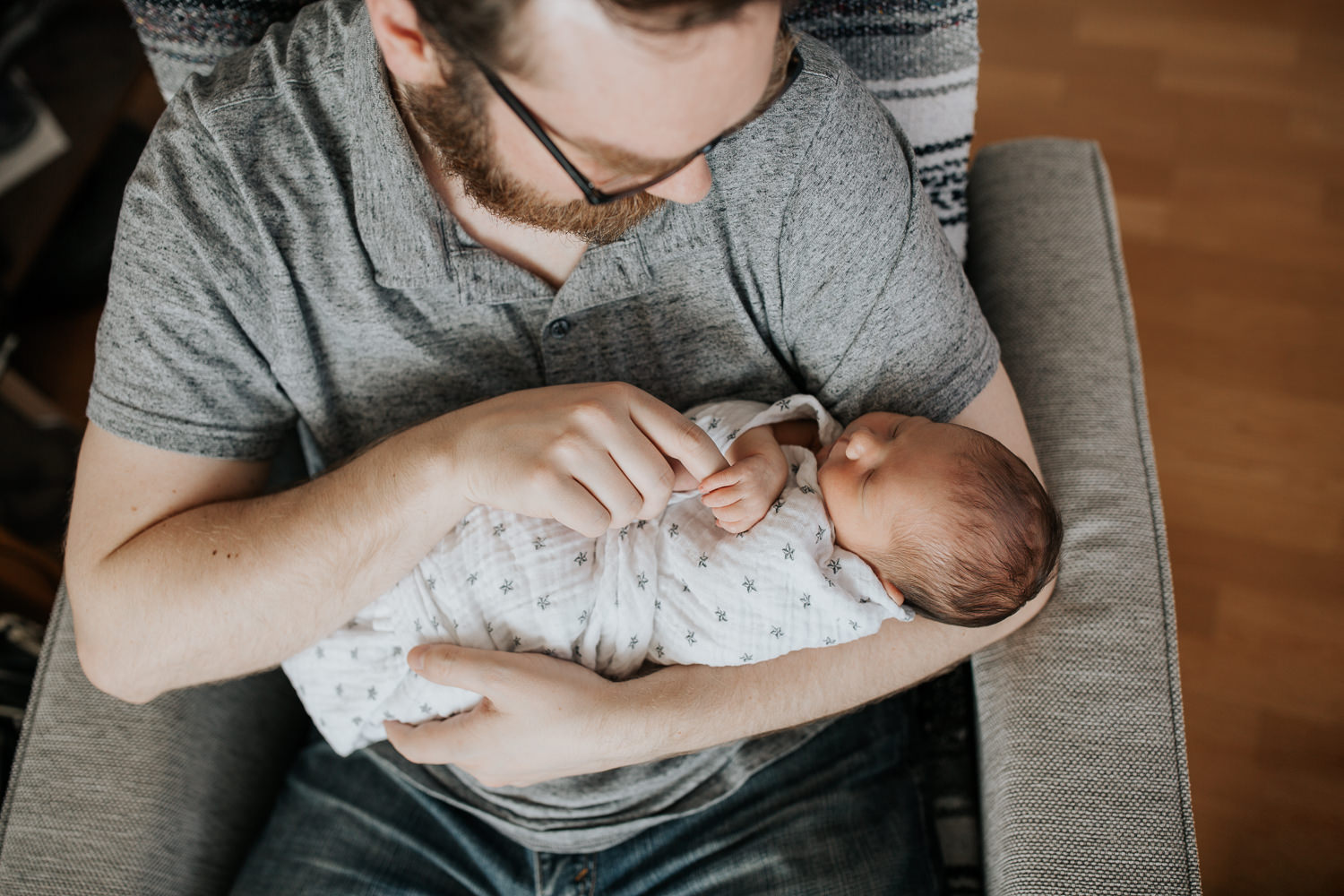 new father sitting in chair holding sleeping 2 week old baby boy in swaddle with dark hair, son holding on to dad's index finger - Markham In-Home Photos