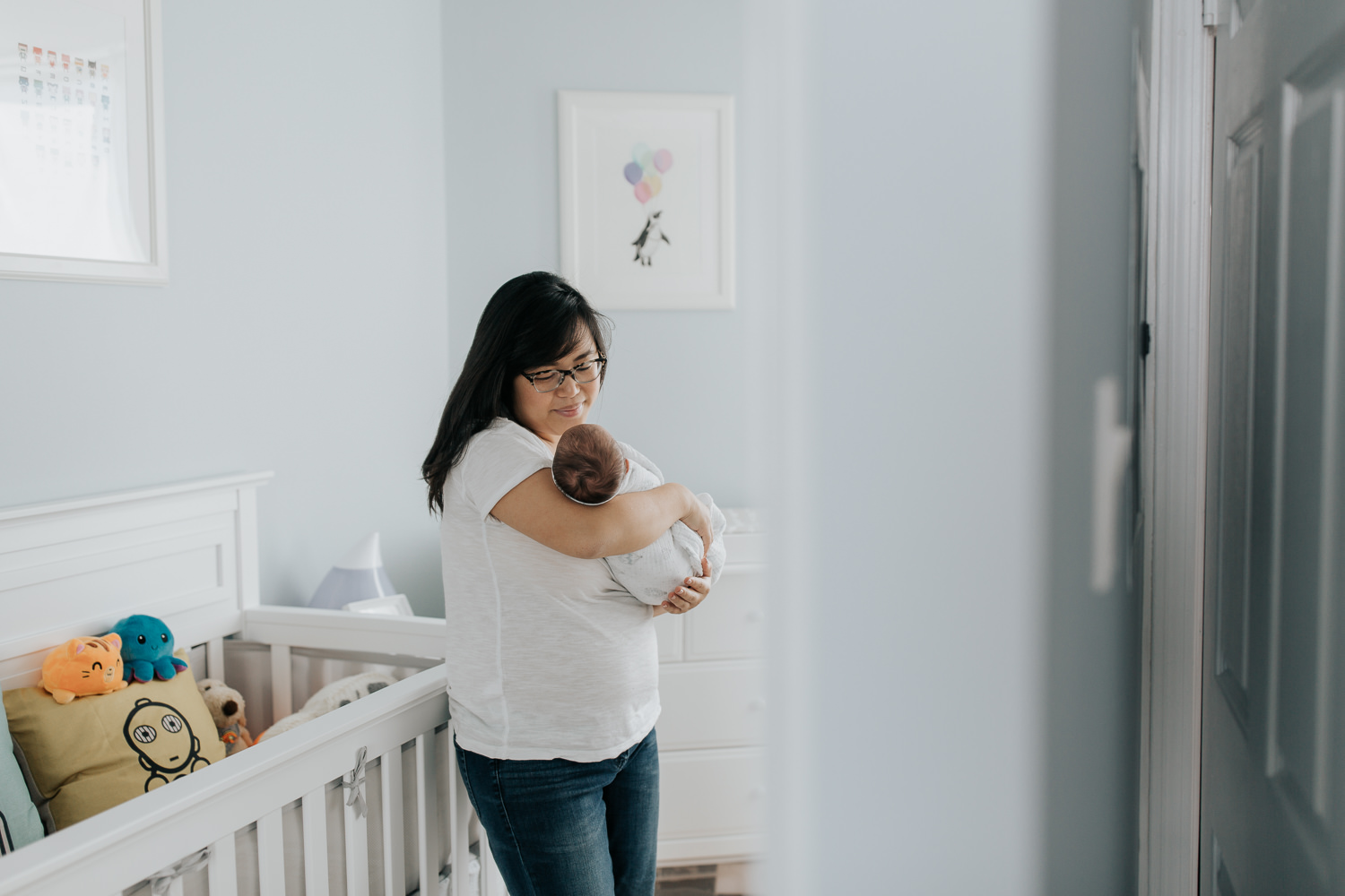 new mother with long dark hair wearing glasses standing in blue nursery holding 2 week old baby boy in her arms, smiling down at son - Barrie Lifestyle Photography