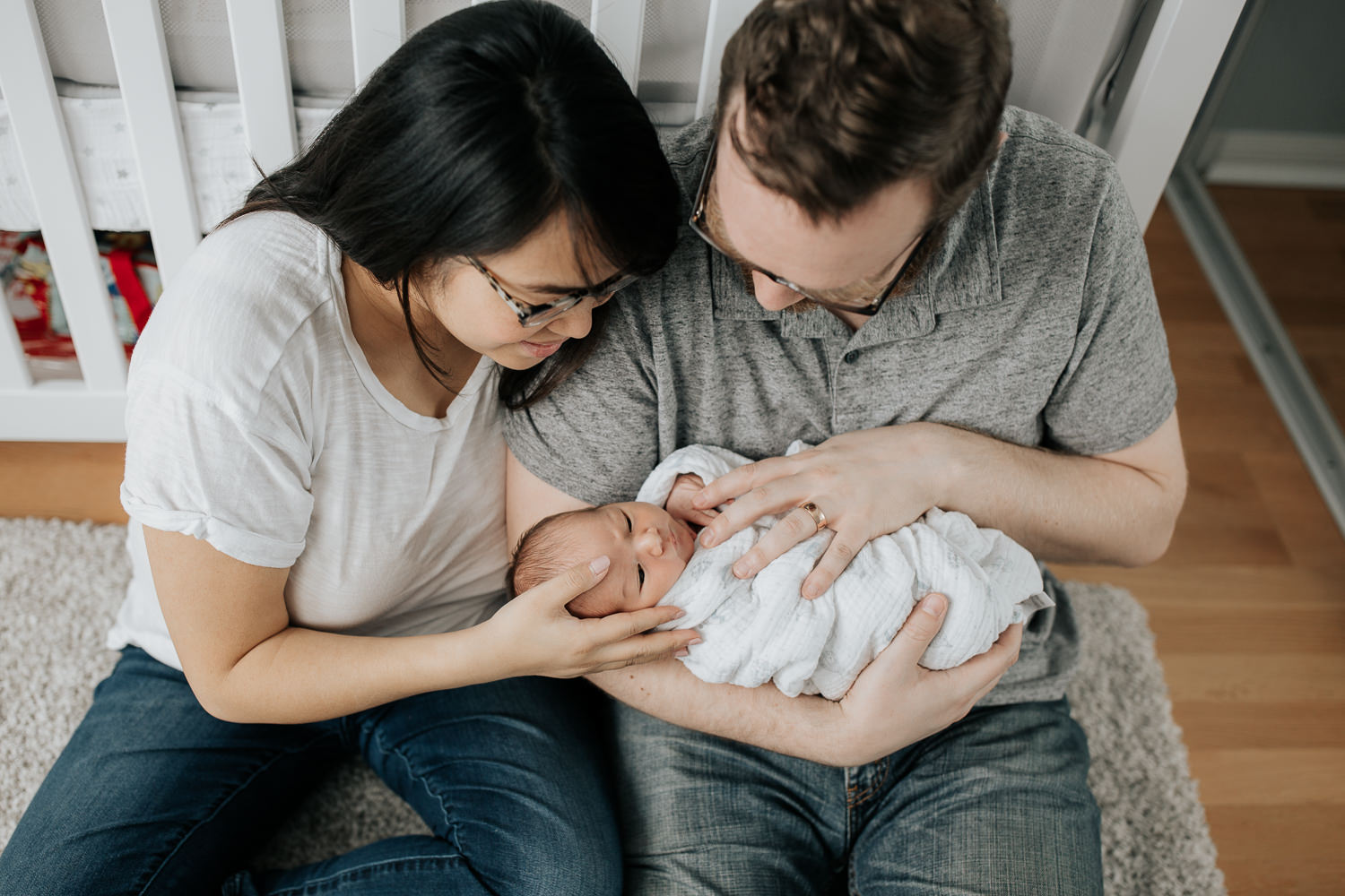 new parents sitting on nursery floor leaning against crib, dad holding 2 week old baby boy in his arms, mom snuggled next to them, looking down at son - Markham In-Home Photography