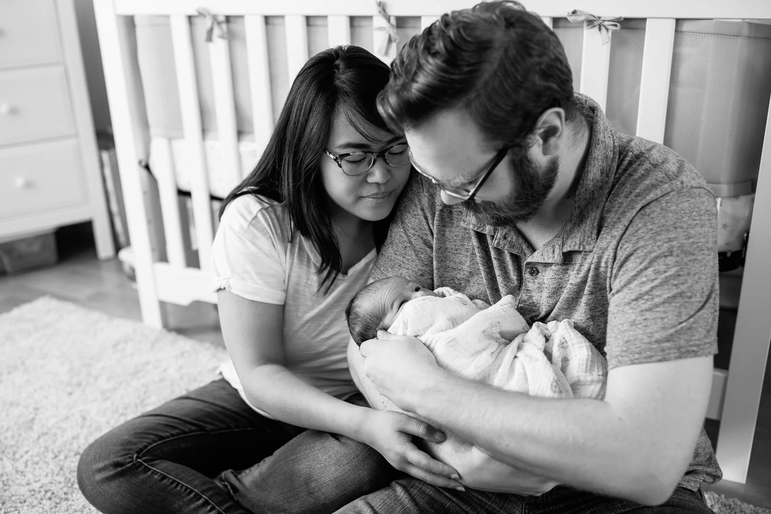 new parents sitting on nursery floor leaning against crib, dad holding 2 week old baby boy in his arms, mom snuggled next to them, looking down at son - Stouffville In-Home Photography