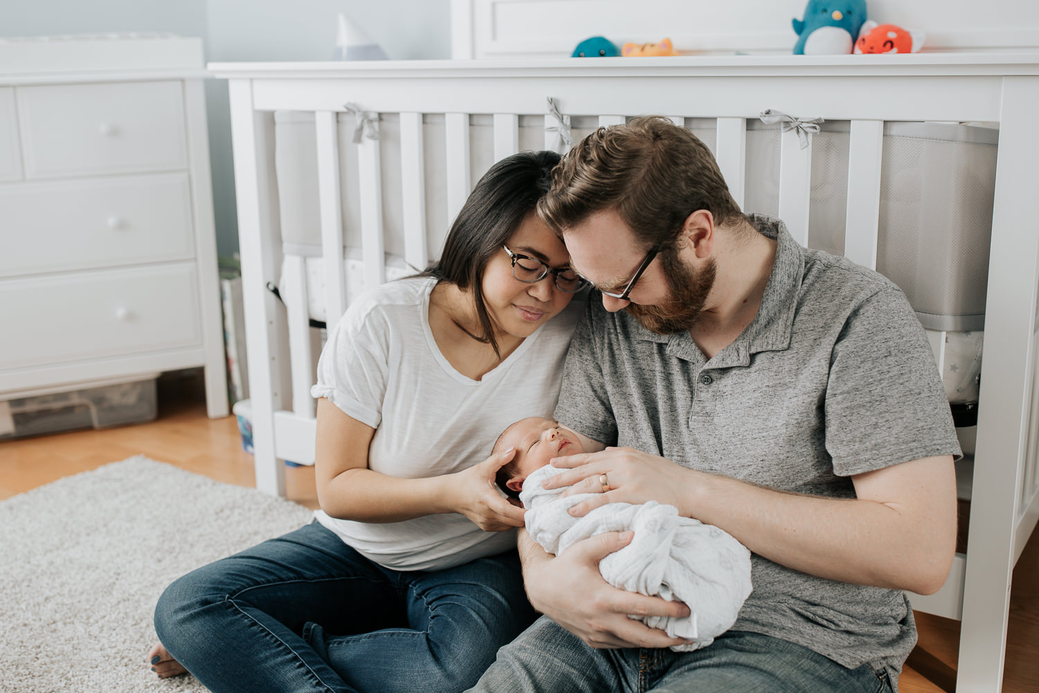 new parents sitting on nursery floor leaning against crib, dad holding 2 week old baby boy in his arms, mom snuggled next to them, looking down at son - Barrie In-Home Photography
