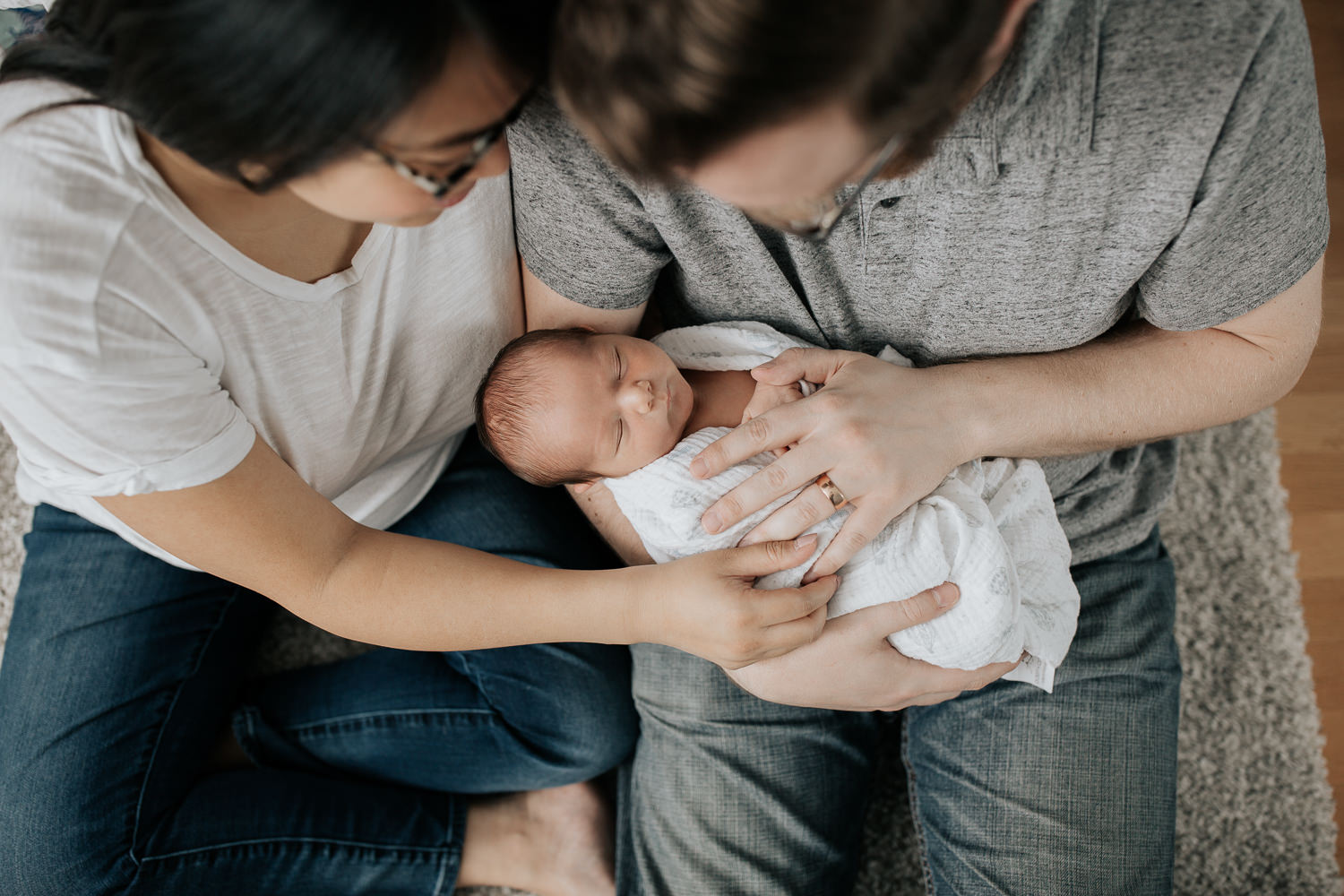 new parents sitting on nursery floor leaning against crib, dad holding 2 week old baby boy in his arms, mom snuggled next to them, looking down at son - GTA In-Home Photography