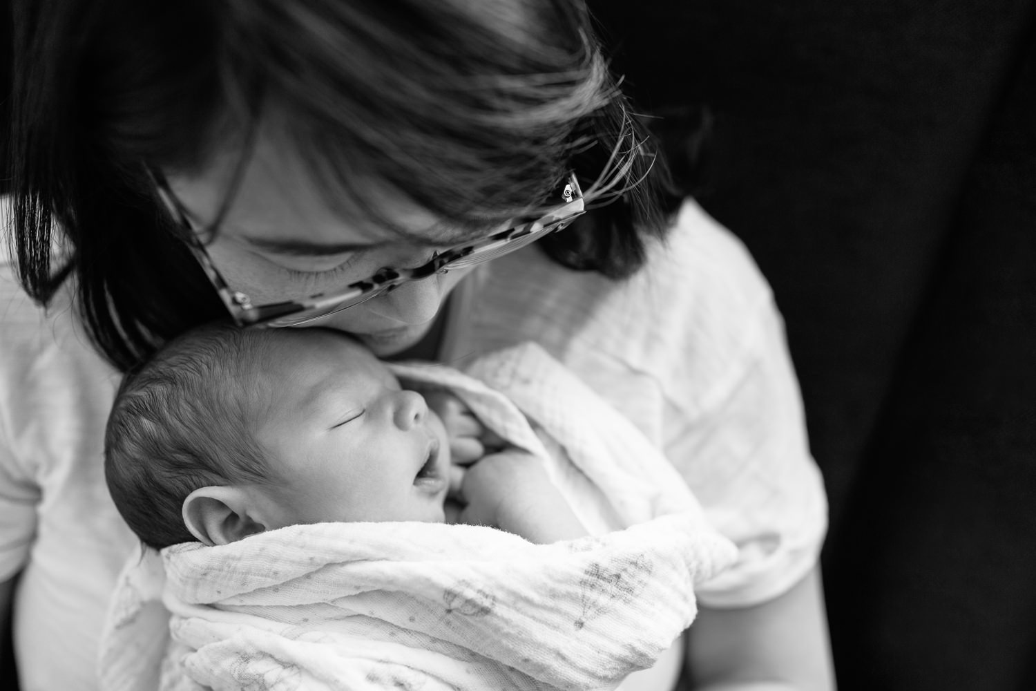 new mother with long dark hair sitting in nursery chair with 2 week old baby boy in swaddle asleep on her chest, mom's cheek resting against top of son's head - Stouffville Lifestyle Photos