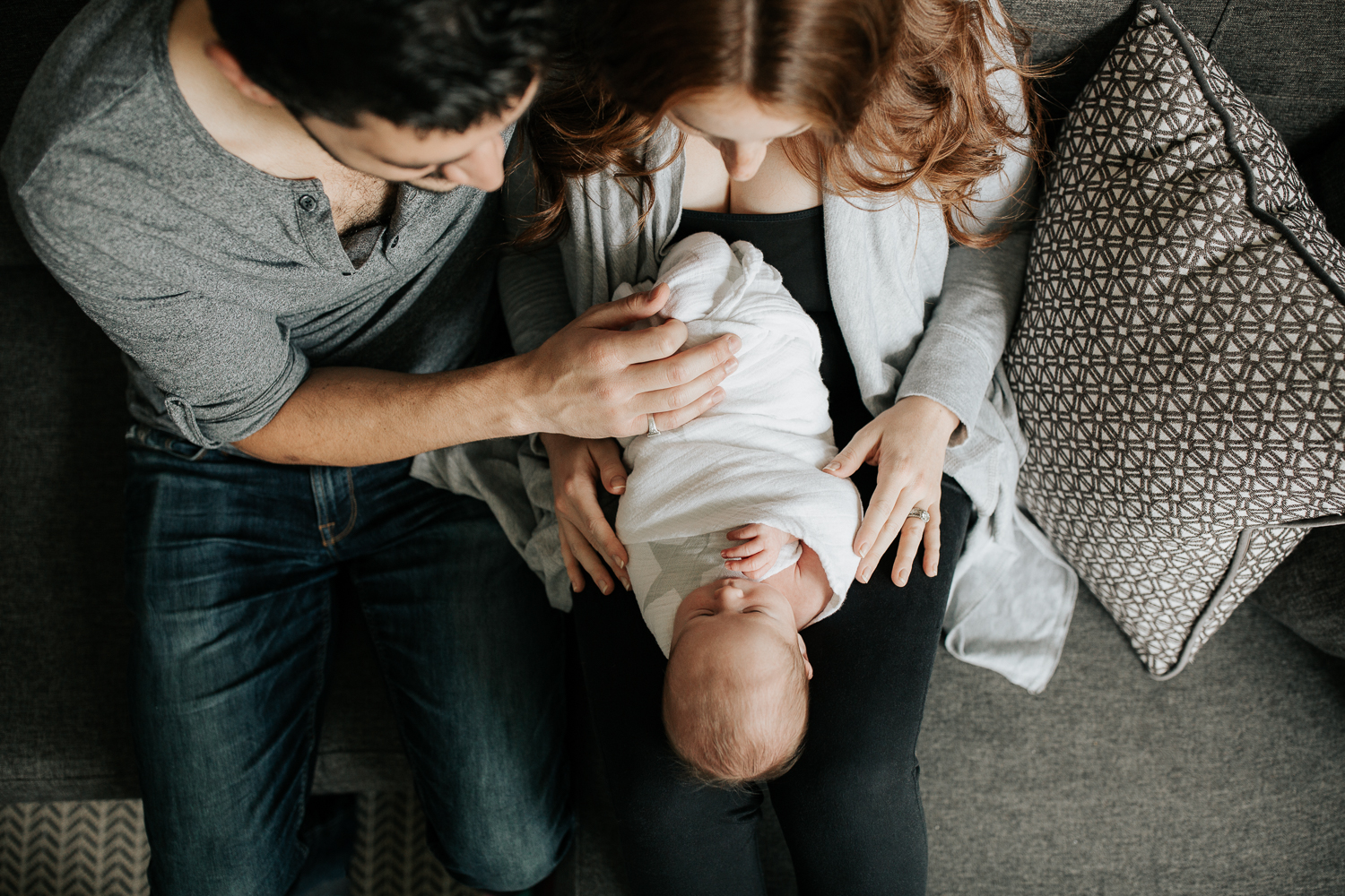 family of 3 sitting on living room couch, 2 week old baby boy lying in mom's lap, dad sitting next to them with hand on son - York Region Lifestyle Photography