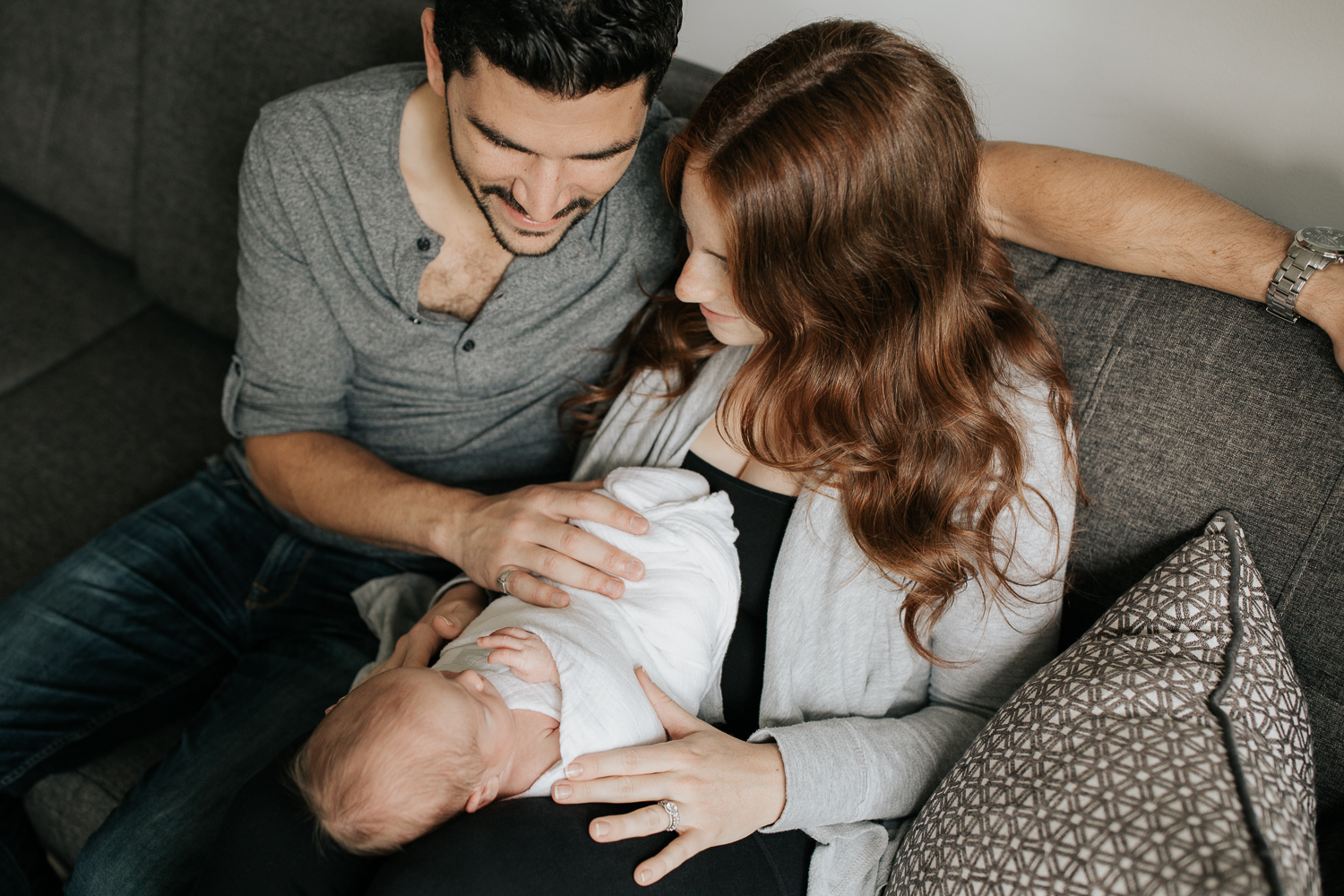 family of 3 sitting on living room couch, 2 week old baby boy lying in mom's lap, dad sitting next to them with hand on son - Newmarket In-Home Photography