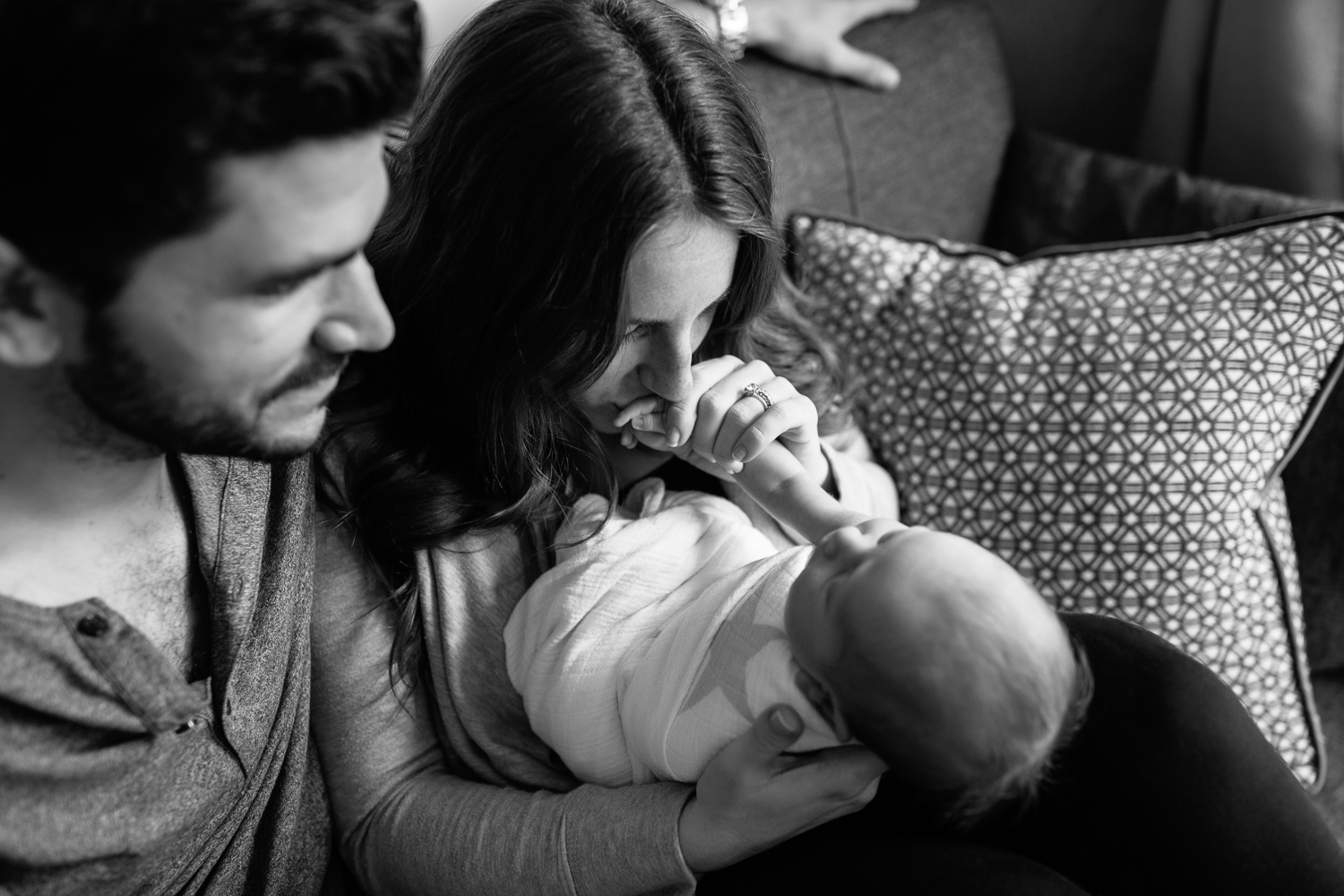 family of 3 sitting on living room couch, 2 week old baby boy lying in mom's lap, dad sitting next to them, mother kissing son's hand - Stouffville In-Home Photography