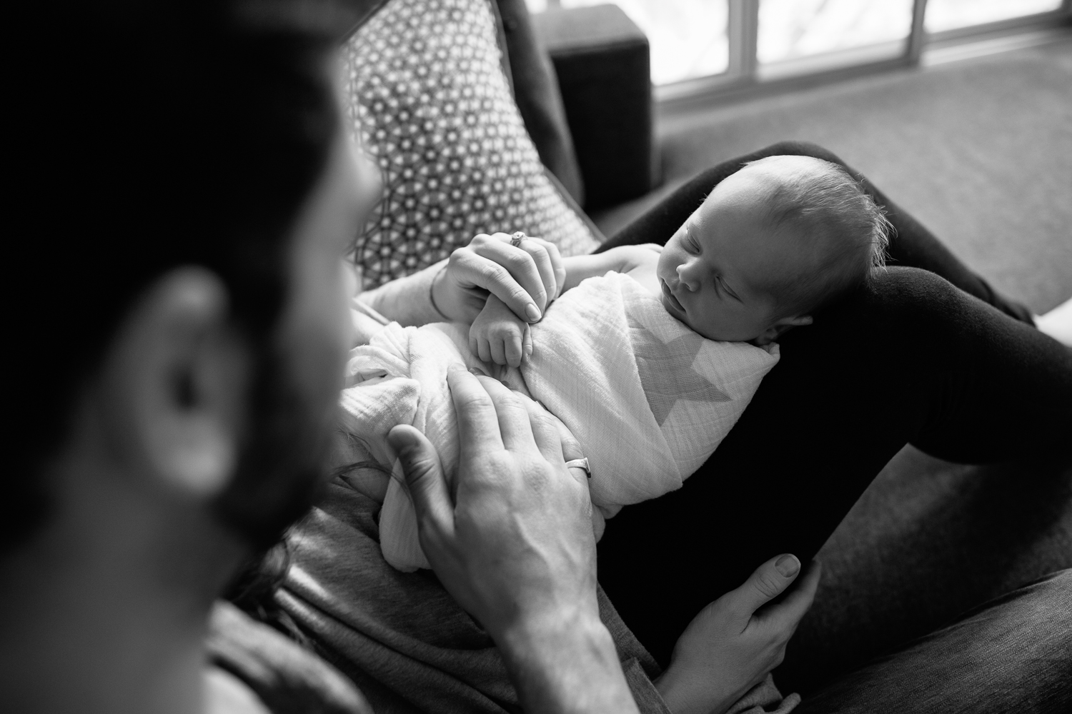 2 week old baby boy lying asleep in mom's lap wrapped in swaddle, mother holding son's arm, father's hand o him - Barrie In-Home Photography