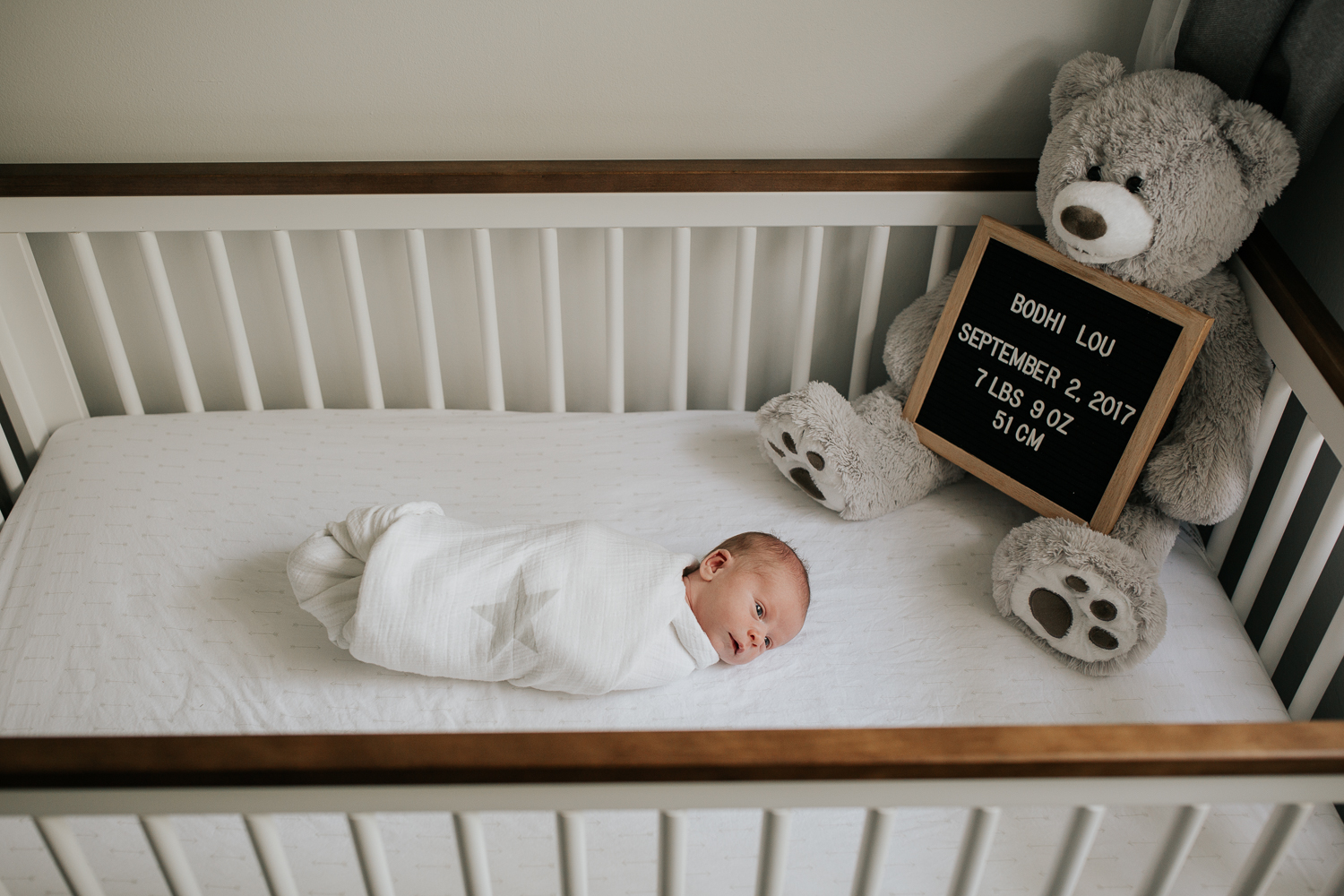 2 week old baby boy in white swaddle with grey stars, lying in white crib with wood trim, awake - GTA In-Home Photography