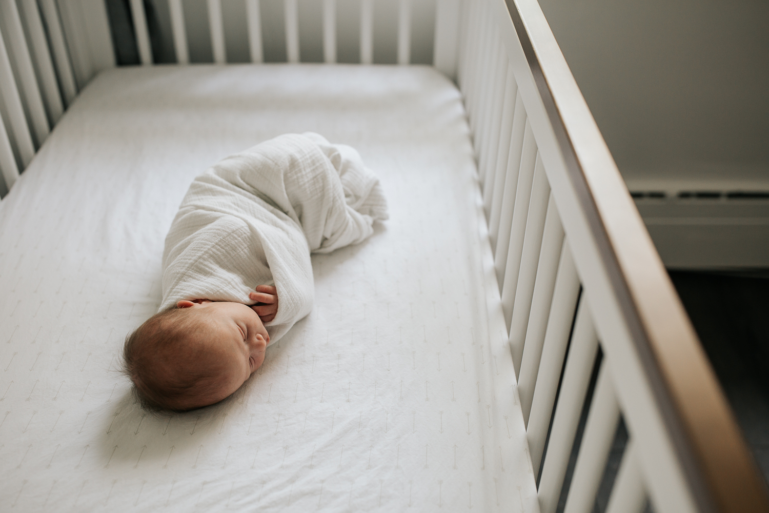 2 week old baby boy in white swaddle with grey stars, lying asleep in white crib with wood trim in neutral nursery - Newmarket In-Home Photos