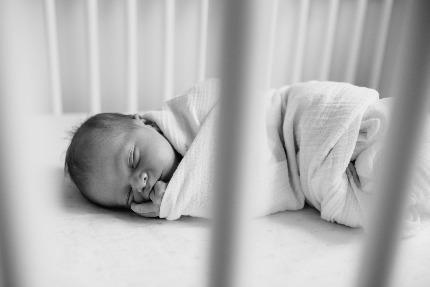 2 week old baby boy in white swaddle with grey stars, lying asleep in white crib with wood trim in nursery, chewing on fingers - Barrie In-Home Photos