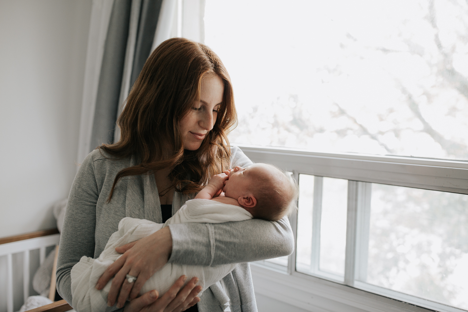 new mom with long red hair standing in neutral nursery holding and snugging 2 week old baby boy wrapped in white swaddle, chewing on hands - GTA In-Home Photos