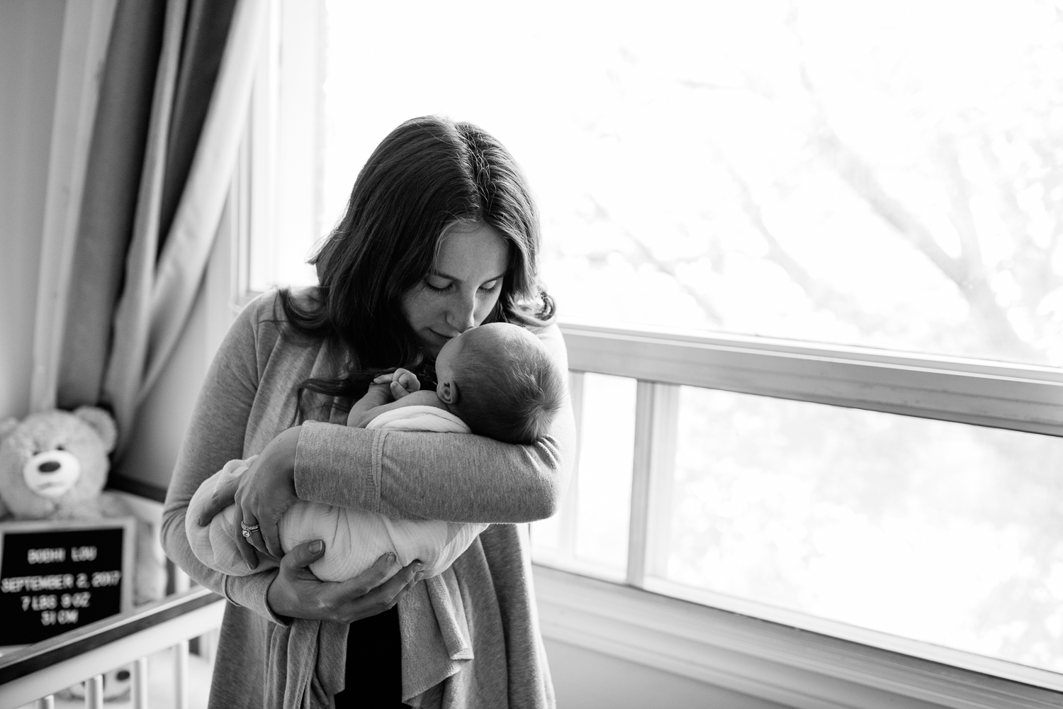 new mom with long red hair standing in neutral nursery holding and snugging 2 week old baby boy wrapped in white swaddle, mother kissing son on head - York Region In-Home Photos