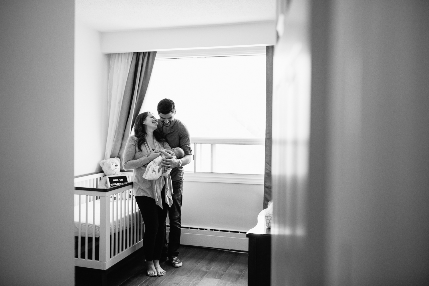 new parents standing in front of crib in neutral nursery, mother holding 2 week old baby boy wrapped in white swaddle, father with arms around wife, hand on son, laughing - Barrie Lifestyle Photos