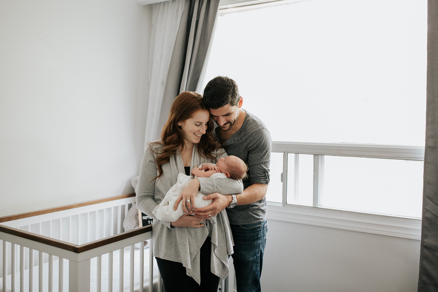 new parents standing in front of crib in neutral nursery, mother holding 2 week old baby boy wrapped in white swaddle, father with arms around wife, hand on son - Markham Lifestyle Photos