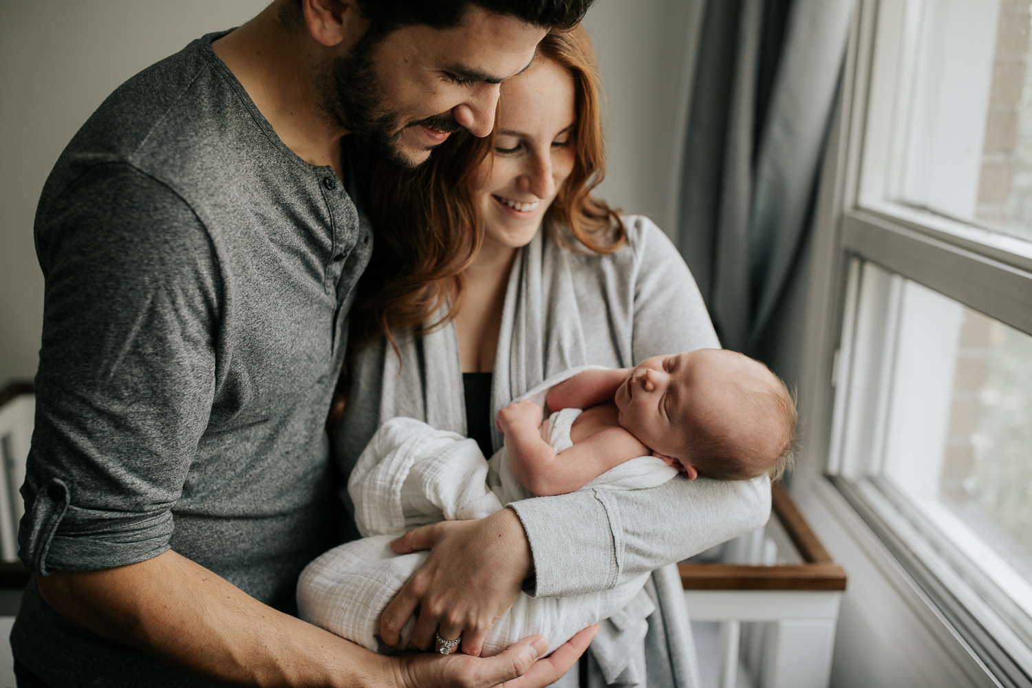 new parents standing in front of crib in neutral nursery, mother holding 2 week old baby boy wrapped in white swaddle, father with arms around wife, hand on son - GTA Lifestyle Photos