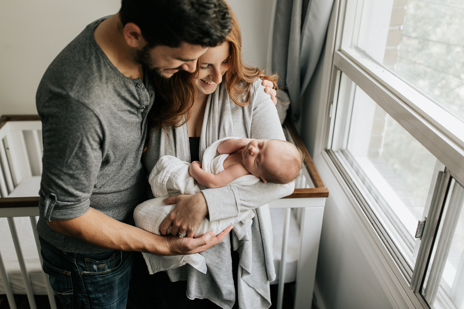 new parents standing in front of crib in neutral nursery, mother holding 2 week old baby boy wrapped in white swaddle, father with arms around wife, hand on son - Stouffville Lifestyle Photography