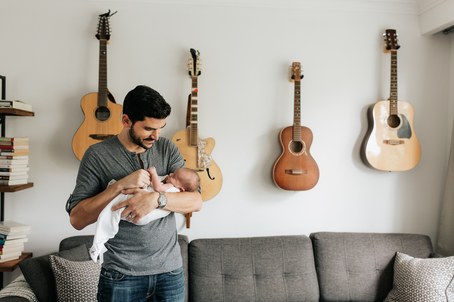 new dad standing in living room in front of wall of guitars, holding 2 week old sleeping baby boy in white swaddle, holding his hands - Barrie Lifestyle Photography